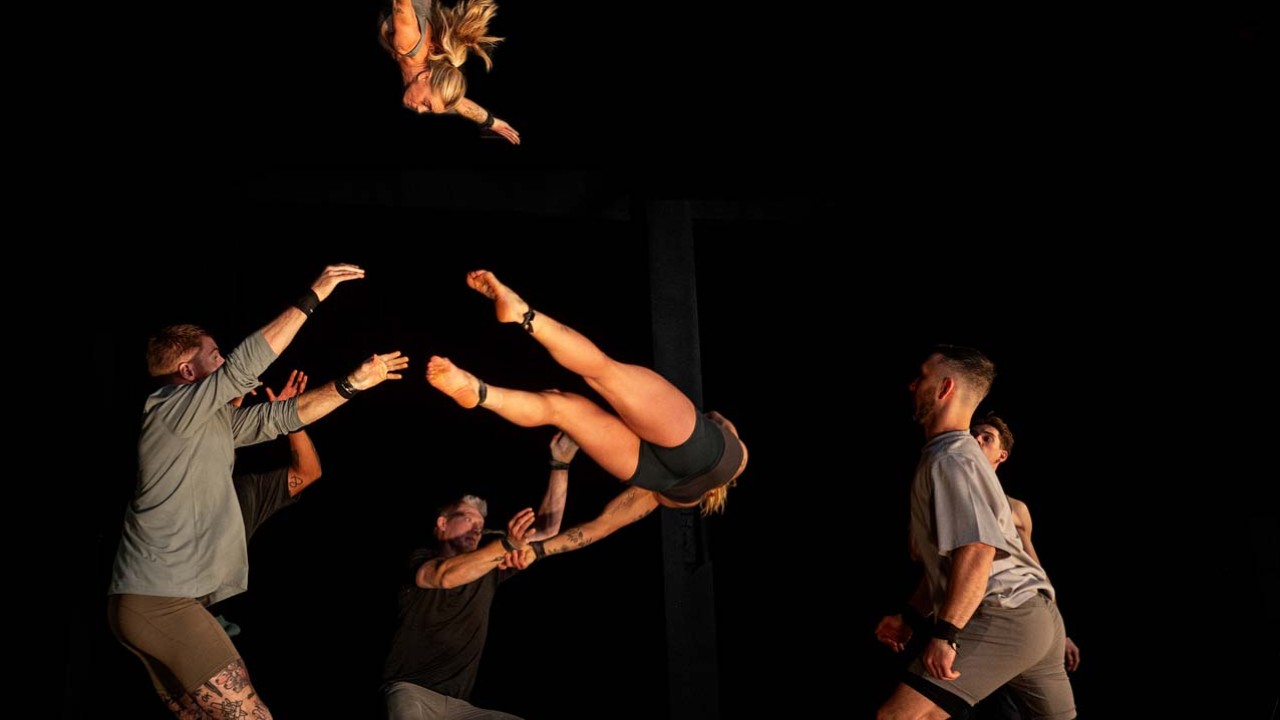 A performer is being elevated in acrobatic poses by a group of dancers in a dark setting.