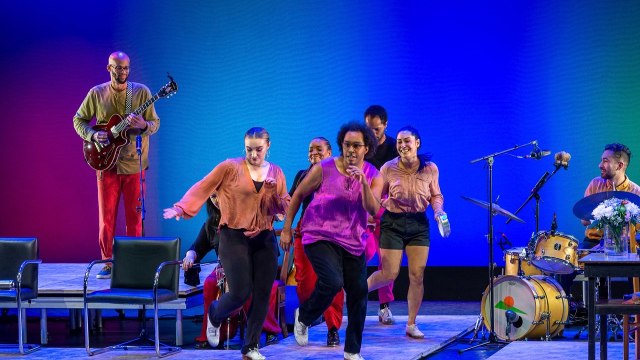 Dancers in bright shirts perform on a blue-green stage with musicians behind them