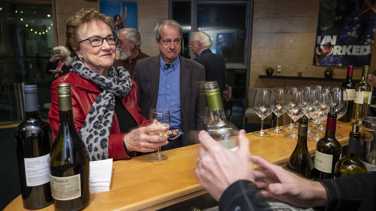 A person in a red jacket enjoys a drink at a bar, with a person in the background and wine bottles visible.