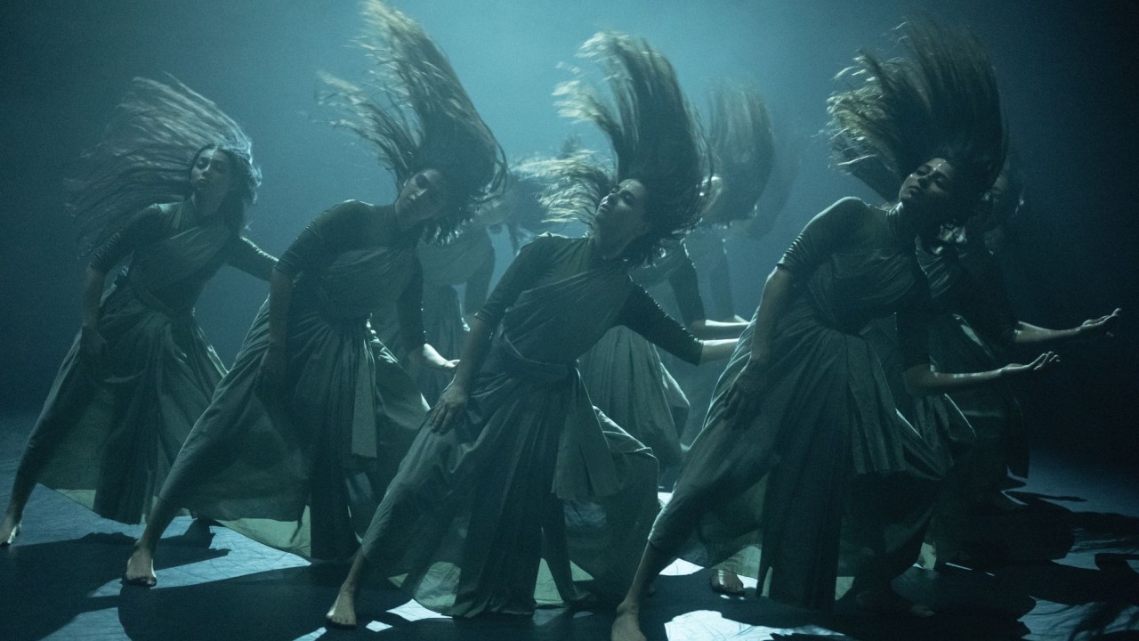Multiple dancers in flowing gowns, hair and fabric mid-swish under moody blue-green stage lighting