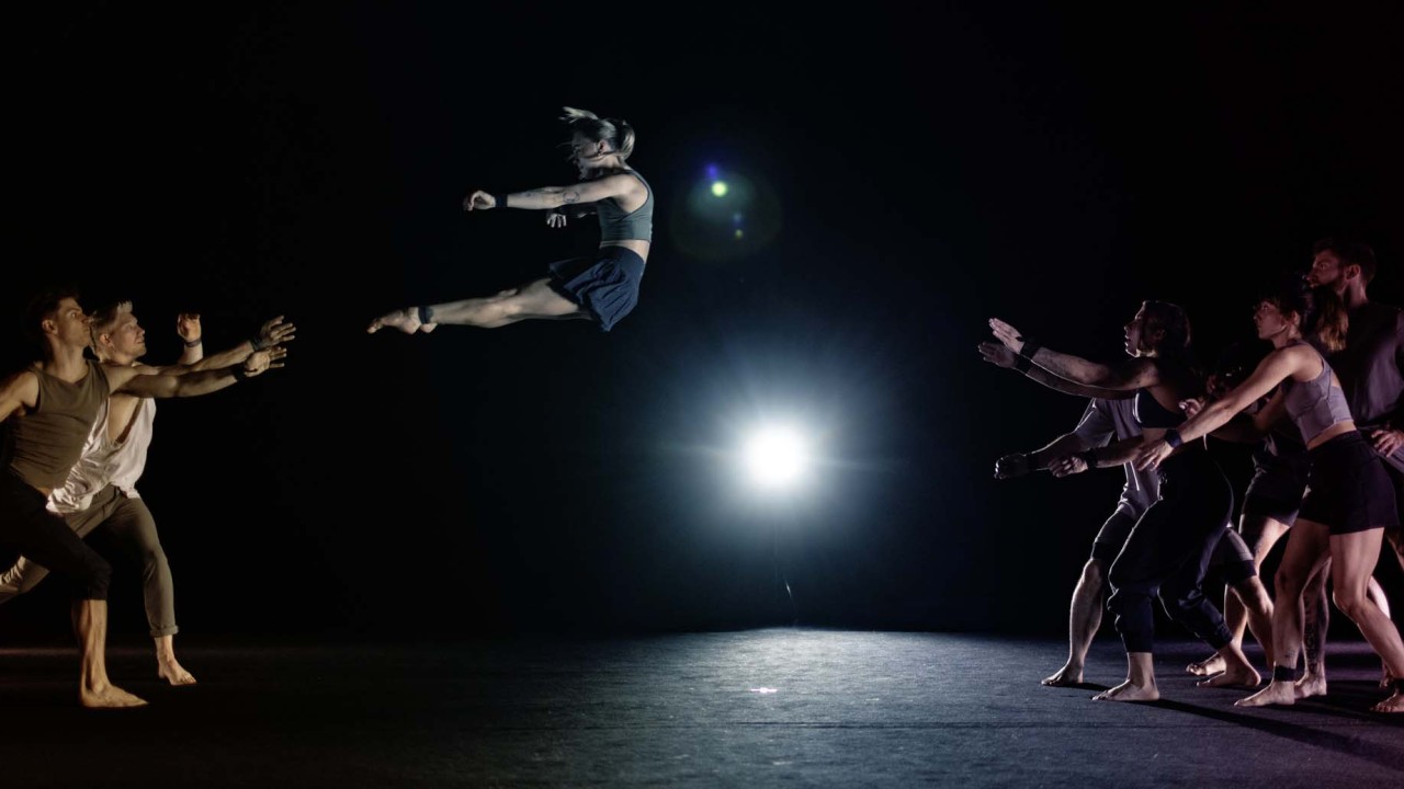 A dancer leaps in mid-air while other performers reach out toward them in a spotlight.