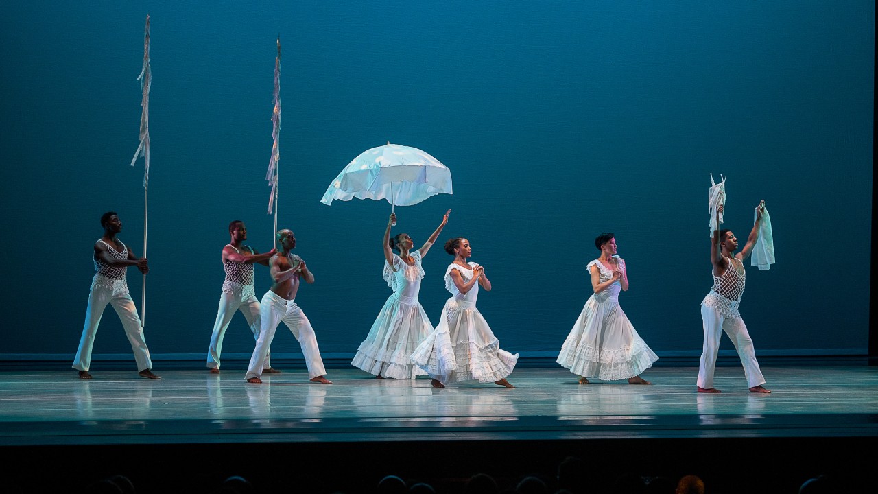 Six dancers in white costumes perform on stage with props including a parasol and poles.