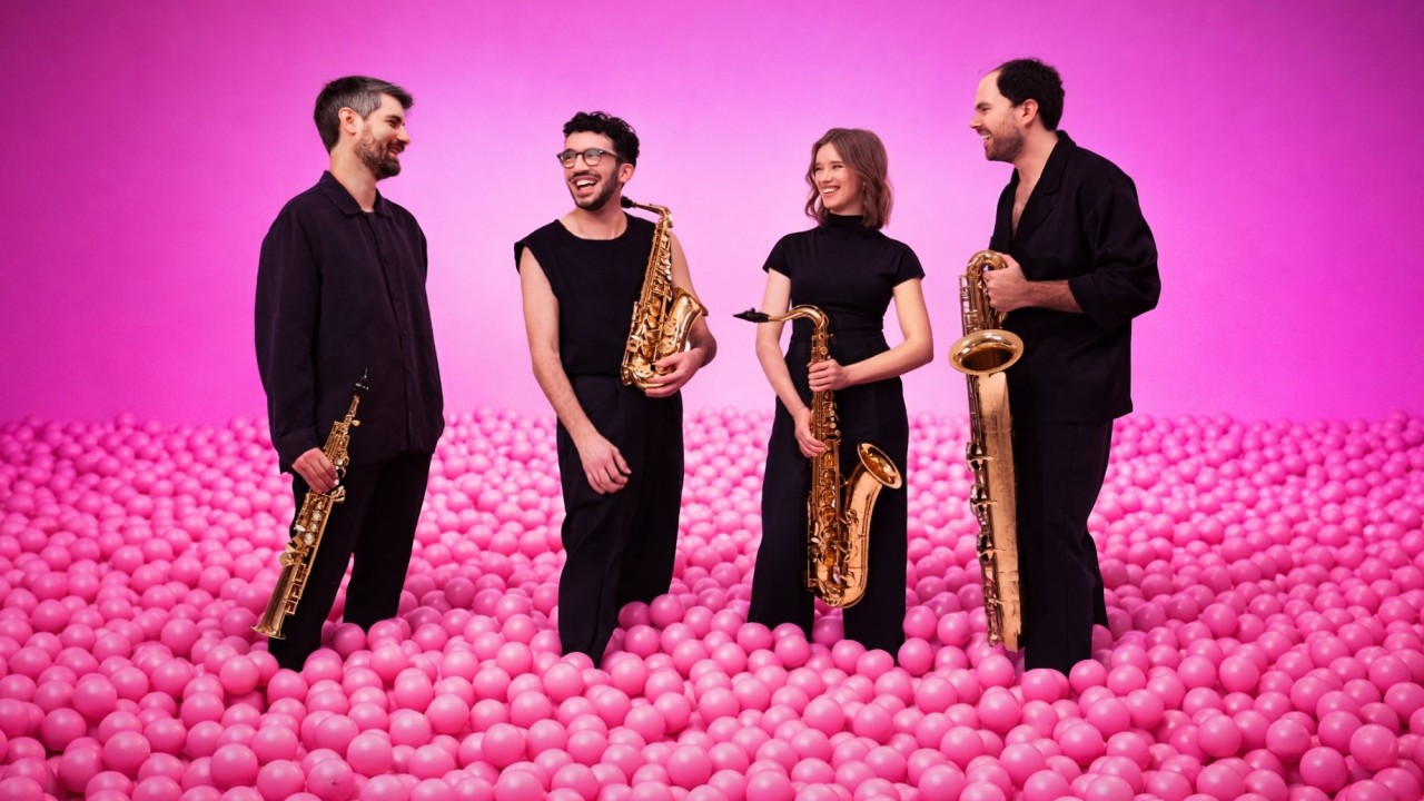 Four saxophonists in black standing in a sea of pink balls against pink backdrop