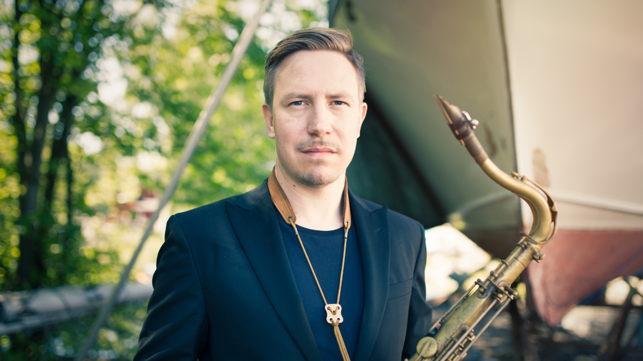 Håkon Kornstad holds a saxophone, standing outdoors near a boat.