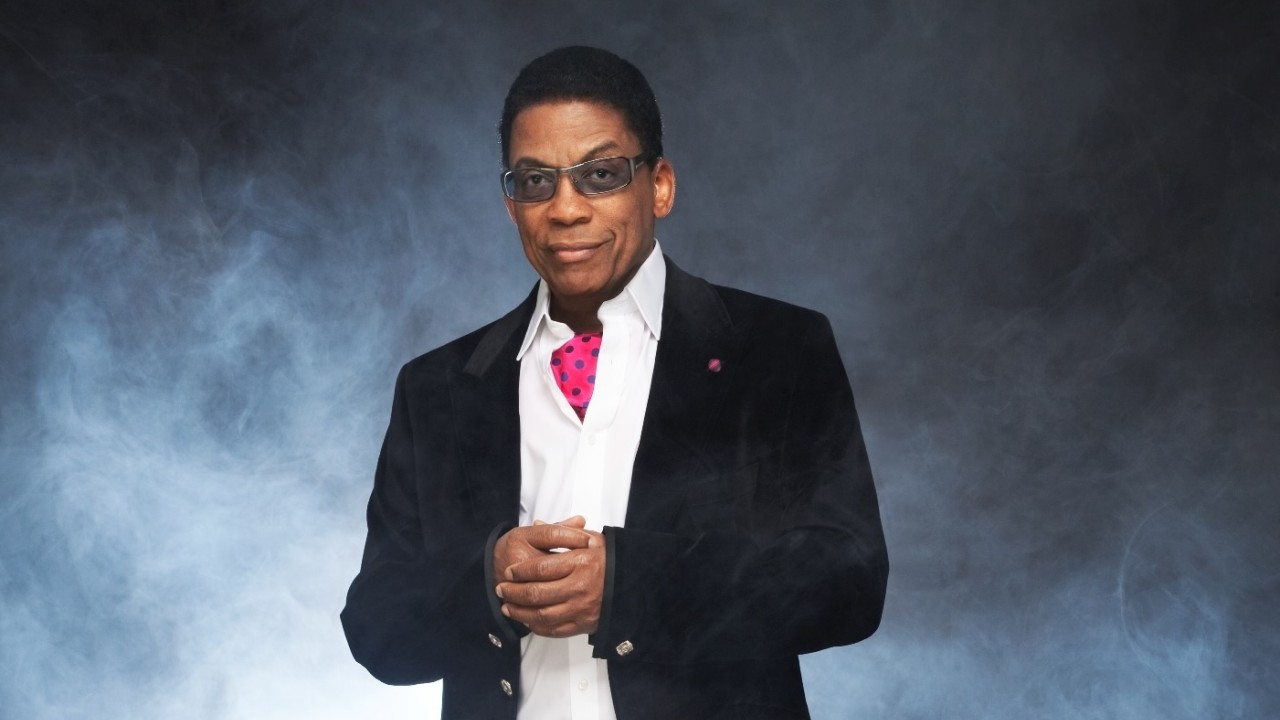 Herbie Hancock in black tuxedo, pink tie and sunglasses against smoky blue background