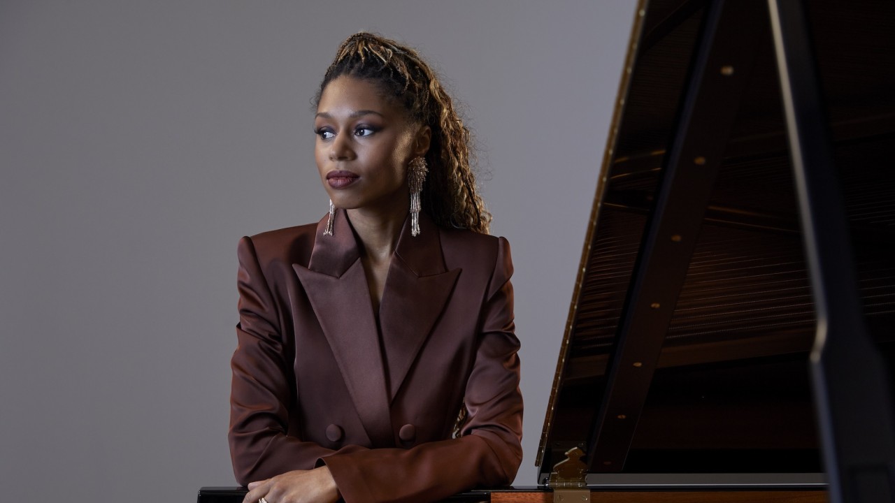 Isata Kanneh Mason in brown blazer with braided ponytail leaning on open grand piano, gazing to the side