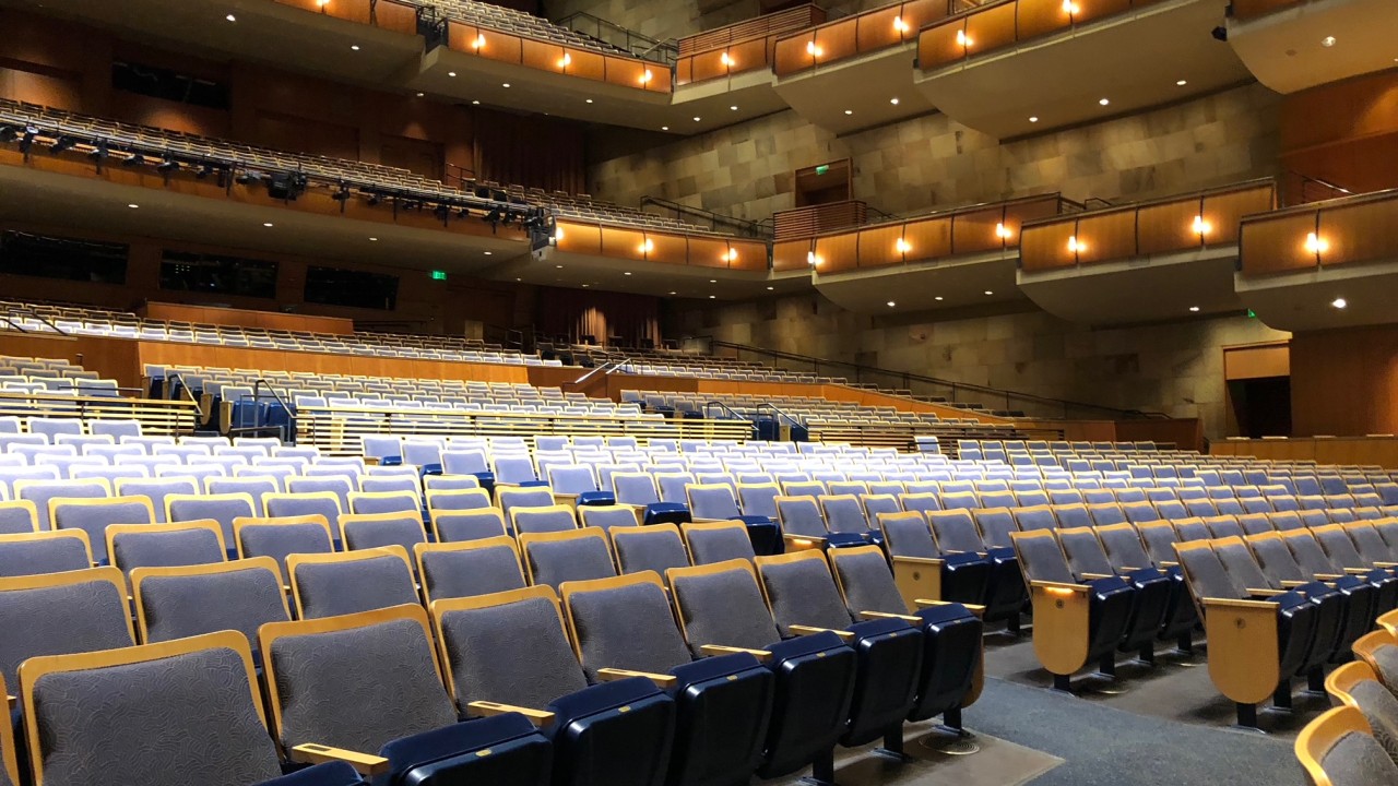Interior view of Jackson Hall,with rows of empty blue and gray seats facing the stage. Multiple balcony levels with warm lighting and curved railings rise above the main floor, highlighting the spacious design of the performance venue before an event begins.