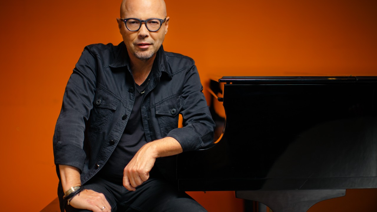 John Beasley in glasses leaning on black grand piano, orange background