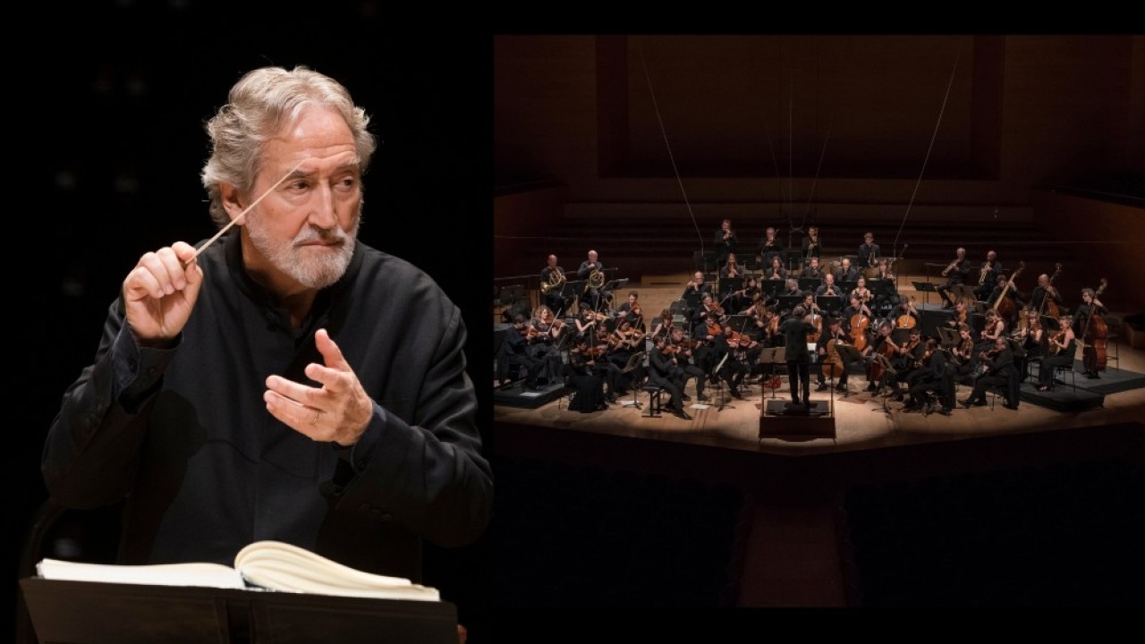 Jordi Savall gesturing beside a full symphony orchestra on stage