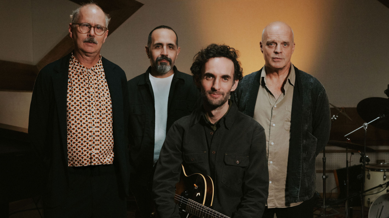 Julian Lage Quartet with John Medeski, Jorge Roeder, & Kenny Wollesen standing behind Julian Lage who is sitting and holding an electric guitar.