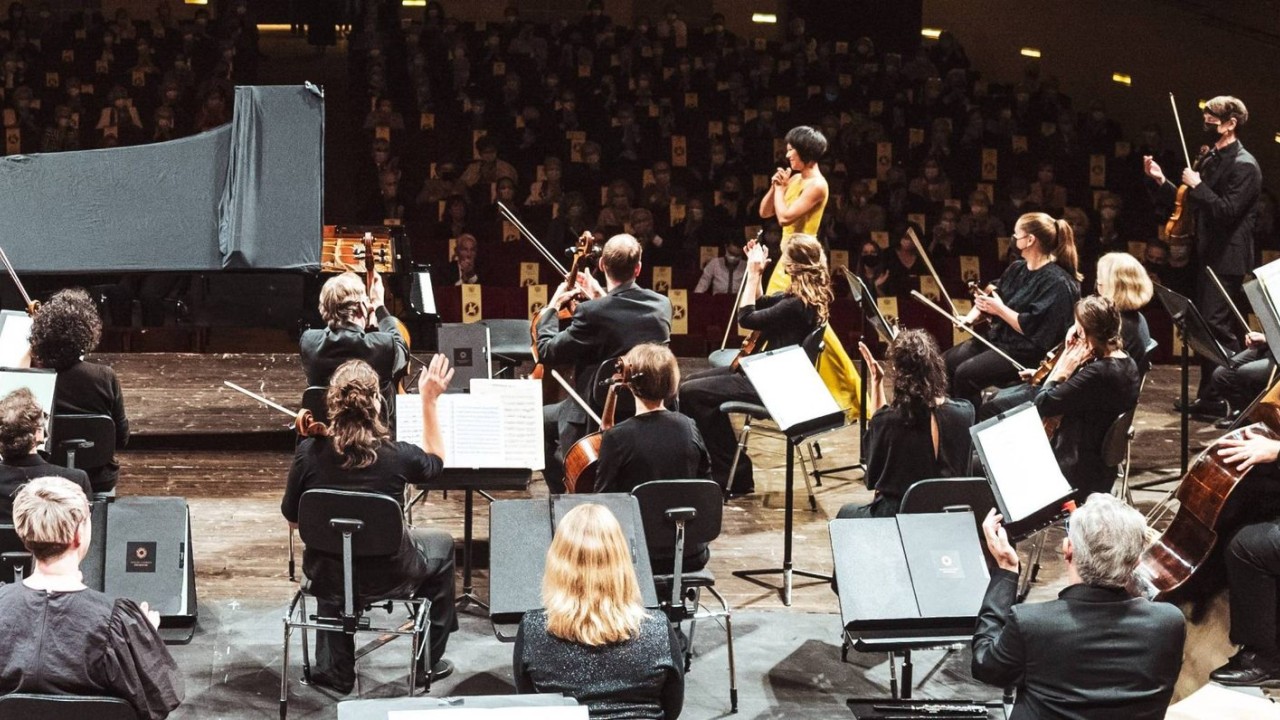 A full orchestra sits on a concert hall stage, applauding a soloist in a bright yellow dress who stands at center near a grand piano. Musicians in formal black attire hold violins, cellos, and other instruments, while a conductor stands to the side. Rows of audience members fill the background, creating a formal classical performance setting.  Is this conversation helpful so far?