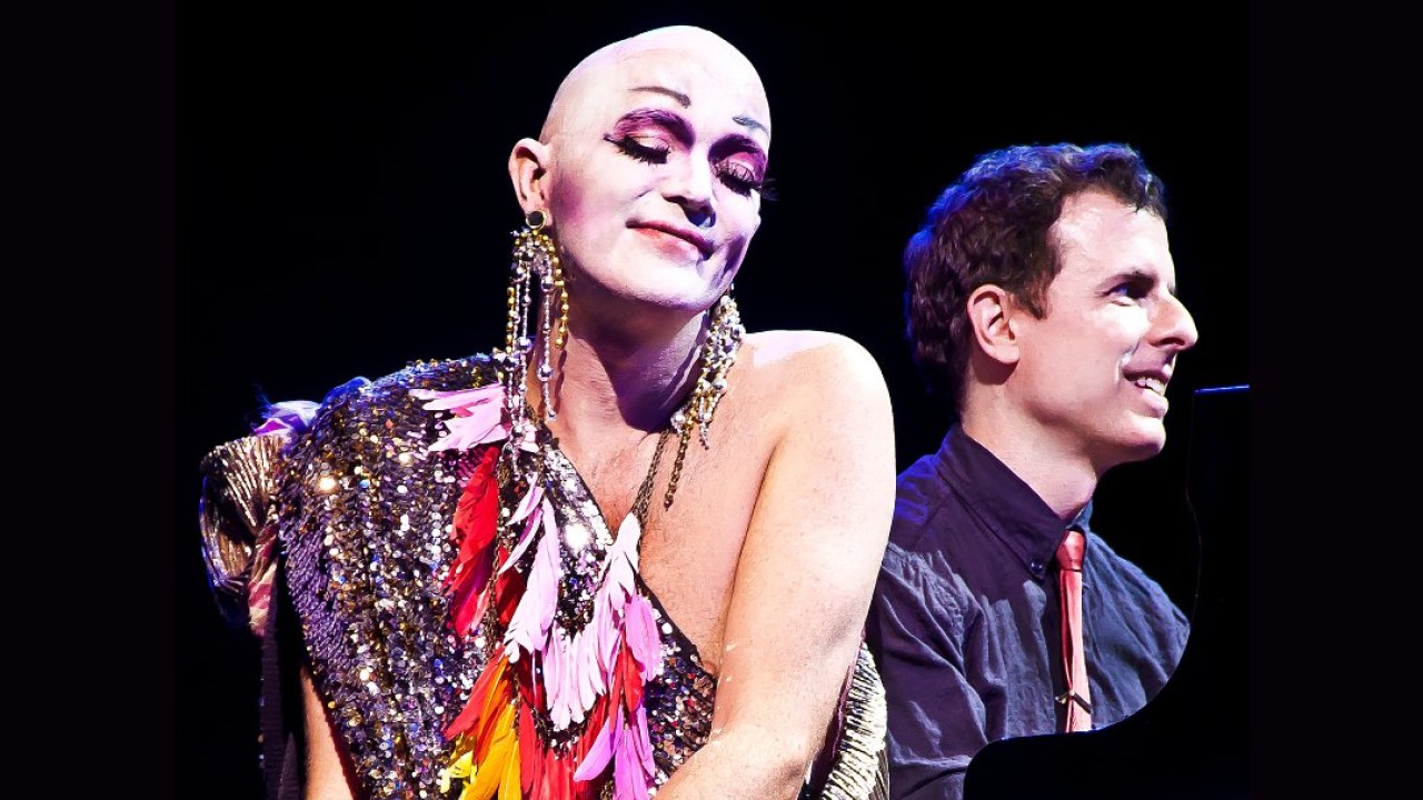 Taylor Mac wears a colorful costume smiles while pianist Matt Ray plays beside them.