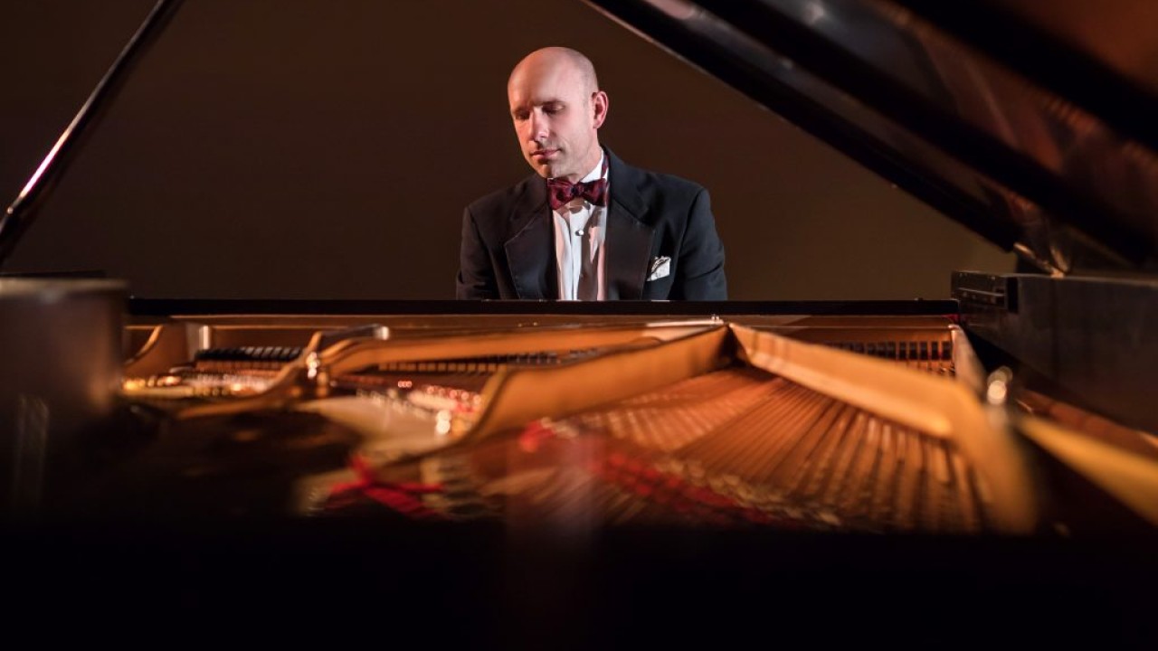 Christopher Taylor in suit and bow tie at grand piano, seen through open piano strings (photo)