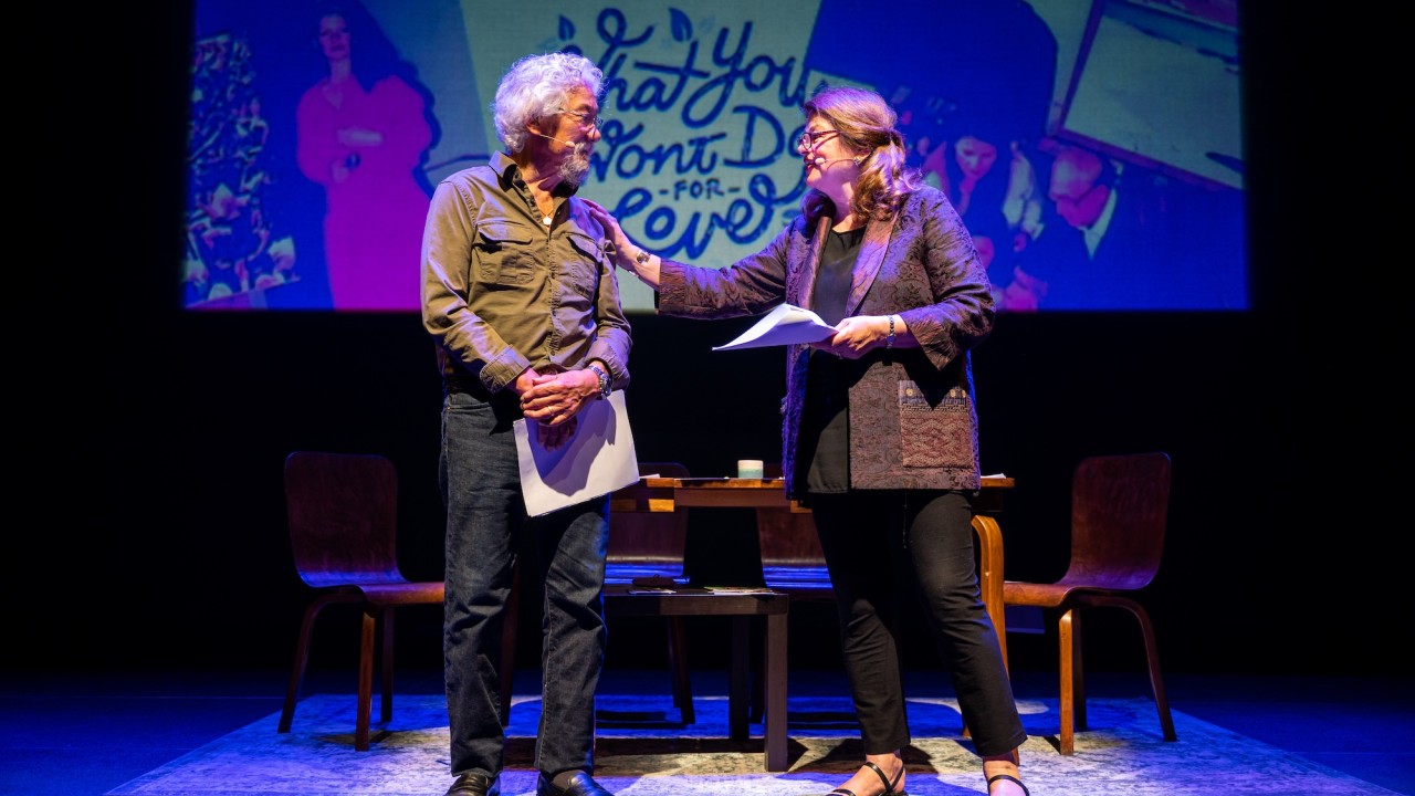 David Suzuki and Tara Cullis facing each other on stage, a colorful projected backdrop and blue stage lighting