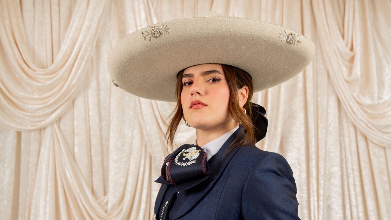 Camila Fernández in white sombrero and navy charro suit, neutral expression before cream drapes