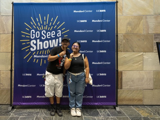 Two people stand smiling in front of a blue “Go See a Show!” Mondavi Center and UC Davis backdrop. They pose playfully using novelty props on sticks, such as glasses and a mustache, suggesting a fun photo opportunity at a campus arts or performance event.