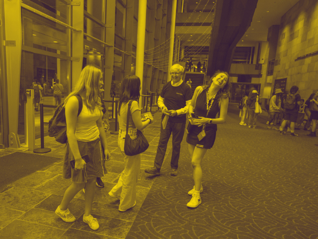 A small group of people chat and laugh inside the Mondavi Center lobby with glass walls and high ceilings. Several young adults stand together holding phones or pamphlets while an older adult speaks with them, suggesting a welcoming or informational moment at a public event or campus venue.