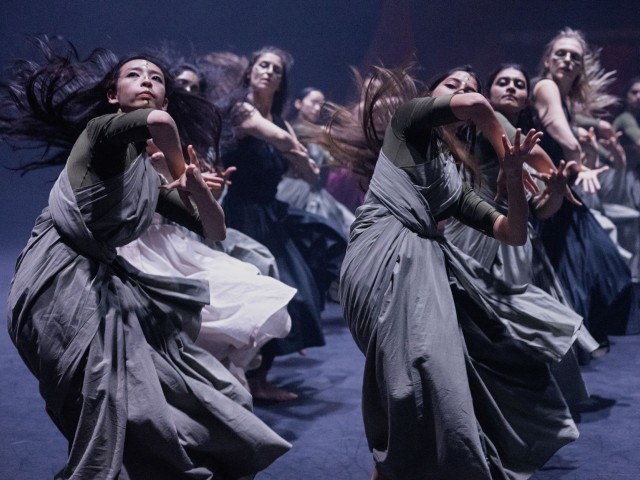 Dancers in flowing gray and white dresses in dynamic contemporary performance on dim stage
