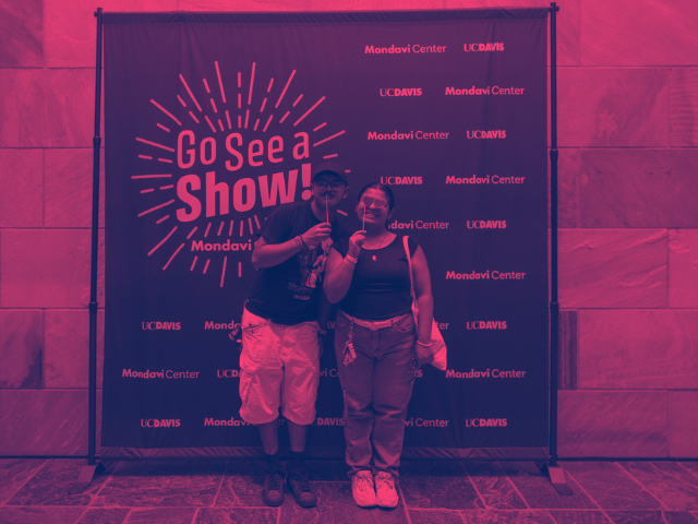 Two people pose together in front of a “Go See a Show!” Mondavi Center and UC Davis step-and-repeat backdrop, tinted with pink and purple lighting. They smile at the camera while holding small novelty props on sticks, suggesting a fun photo moment at an arts or performance event.