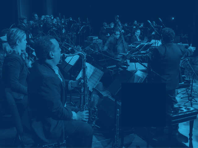 A large jazz orchestra performs on a dimly lit stage with blue and red lighting. Seated musicians play saxophones, trumpets, trombones, piano, bass, and drums while a conductor stands at front right, gesturing toward the ensemble. Music stands and microphones fill the stage, conveying a formal live concert performance.