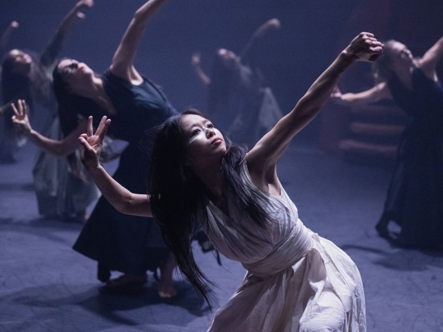 Central dancer in white with face paint reaching upward amid dark-clad ensemble
