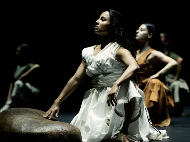 Dancer in white gown kneels in spotlight on dark stage, hand on low prop; two dancers behind
