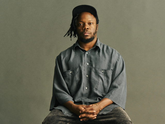 Portrait of Ambrose Akinmusire wearing a lose grey shirt seated against a neutral background.