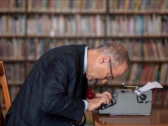 Man in suit hunched over a typewriter with a pipe in his mouth at a library table, bookshelves blurred behind