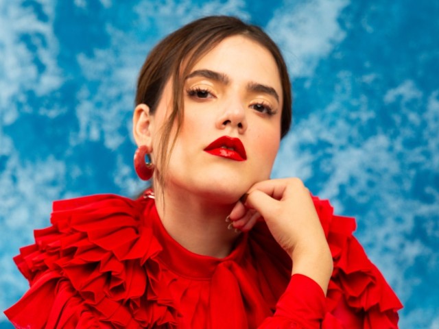 Camila Fernández posed with hand on chin, red ruffled blouse, and a blue backdrop.