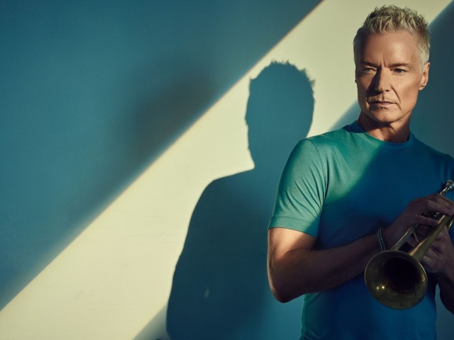 Portrait of an older man holding a trumpet, teal shirt, dramatic shadow.