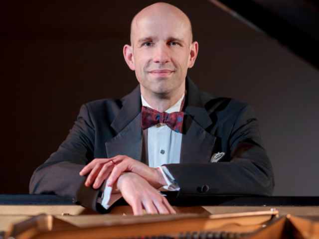 Christopher Taylor in a tuxedo leans on a grand piano with his hands crossed.