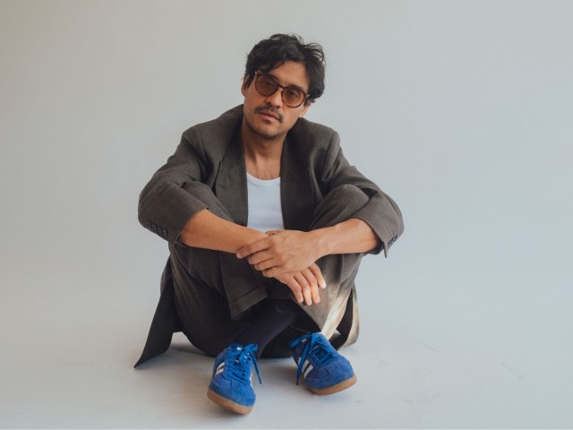 Young Man in blazer and sunglasses sitting cross-legged on plain studio floor, blue sneakers.