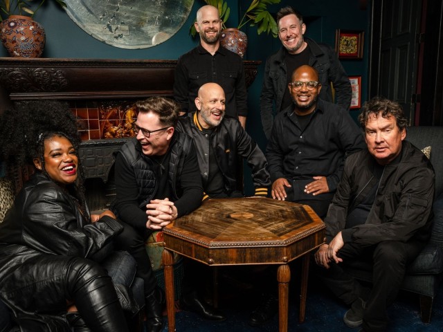 Group portrait of seven people gathered around a small wooden table in a dark cozy room