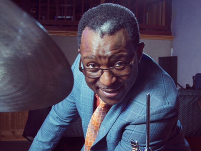 Herlin Riley in a blue suit and an orange tie leans by a drum cymbal