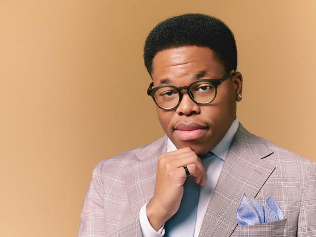 Isaiah J. Thompson smirking at the camera in a tan suit and glasses, hand on chin, in front of a beige background