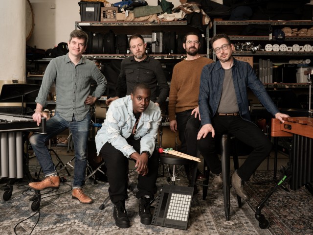 Five musicians pose in a music studio with various instruments and equipment in the background.