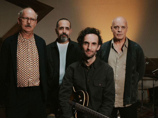 Julian Lage Quartet with John Medeski, Jorge Roeder, & Kenny Wollesen standing behind Julian Lage who is sitting and holding an electric guitar.