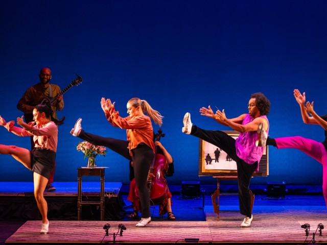 Four dancers mid-kick on blue-lit stage in colorful outfits, with a guitarist and cellist behind.