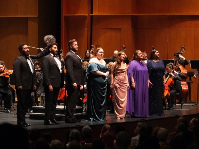 A group of six vocal performers on stage, dressed elegantly, with an orchestra in the background.