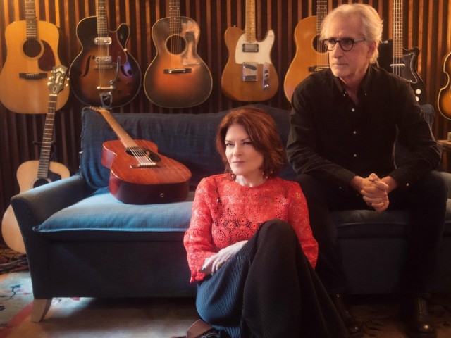Rosanne Cash sits in front of John Leventhal on blue couch in guitar-filled studio