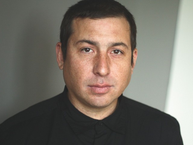 Headshot portrait of Tommy Orange with short dark hair, neutral expression, wearing dark shirt.