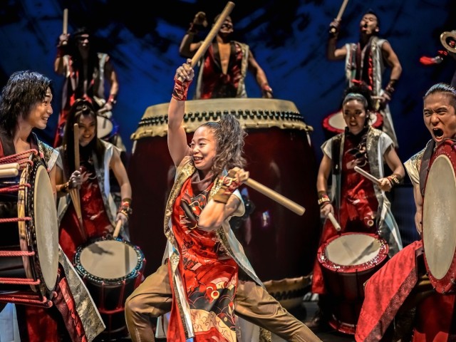 Energetic taiko drummers in red costumes performing with large drums on stage.