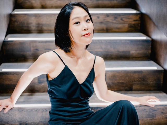 Woman in dark green dress seated on wooden stairs, relaxed posture, direct gaze.