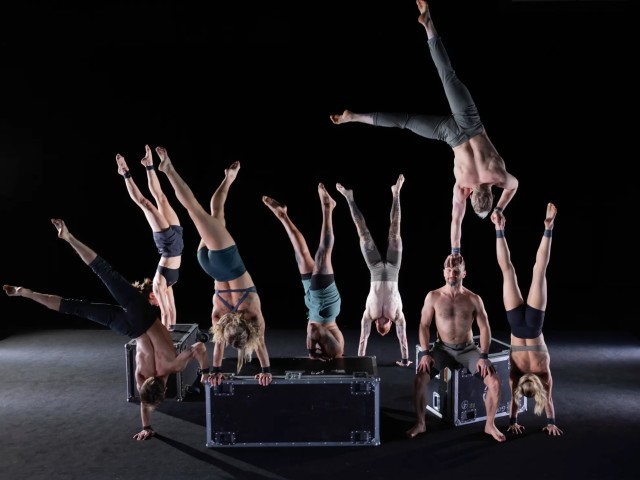 Acrobats forming handstands and midair flips above a box on a dark stage