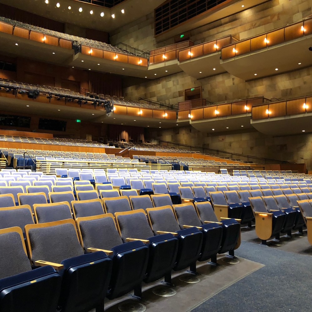 Interior view of Jackson Hall,with rows of empty blue and gray seats facing the stage. Multiple balcony levels with warm lighting and curved railings rise above the main floor, highlighting the spacious design of the performance venue before an event begins.