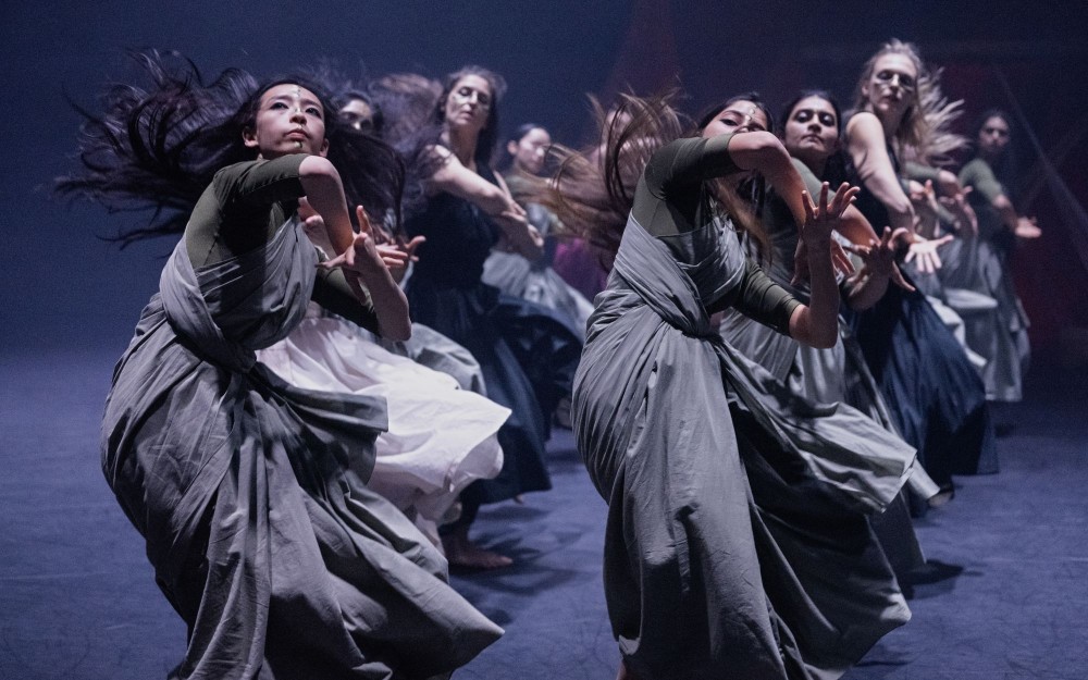 Dancers in flowing gray and white dresses in dynamic contemporary performance on dim stage