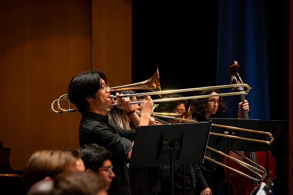 Trombonists in black performing with brass trombones on a wood-paneled stage