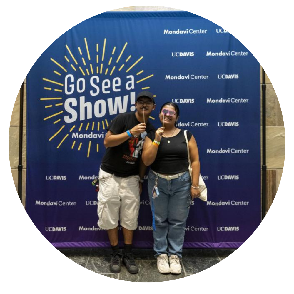 Two people pose together in front of a blue backdrop with "Go See a Show!" text.