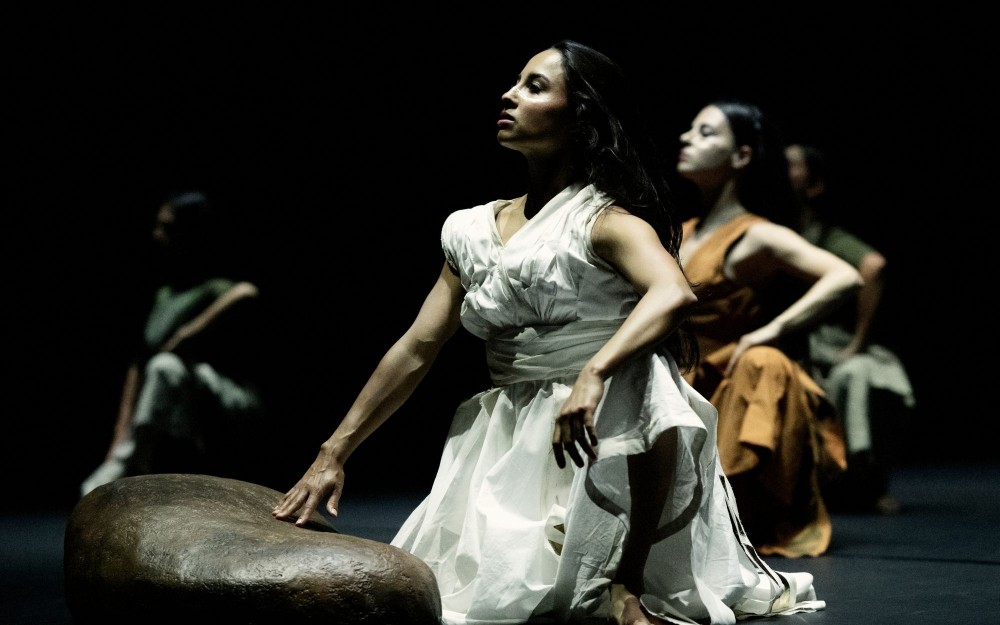 Dancer in white gown kneels in spotlight on dark stage, hand on low prop; two dancers behind
