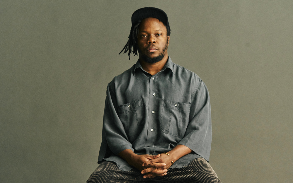 Portrait of Ambrose Akinmusire wearing a lose grey shirt seated against a neutral background.